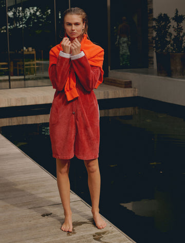 Een vrouw die naast een zwembad staat met een roze badjas aan en een oranje trui om haar nek geslagen