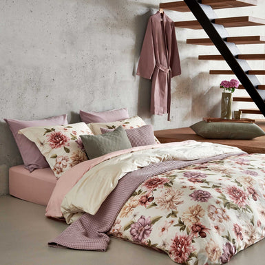 Een bed in een rustige setting met een dekbedovertrek met rode, paarse en beige bloemen. Op het bed ligt een sprei in een lila kleur en in een groene kleur