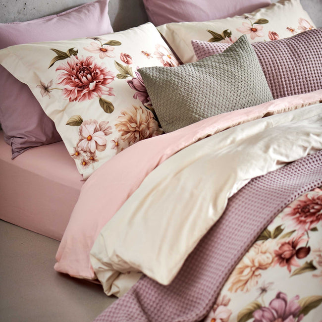 detail shot van een dekbedovertrek in een beige kleur met roze, paarse en beige bloemen op het bed ligt een sprei en een sierkussen in een paarse kleur en een sierkussen in een groene thyme kleur