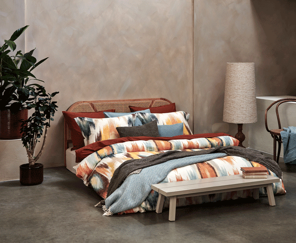 een bed met een dekbedovertrek met een print in oranje en blauwe kleuren met daaronder een dekbedovertrek in een roest bruine/oranje kleur en een blauw wol plaid over het bed